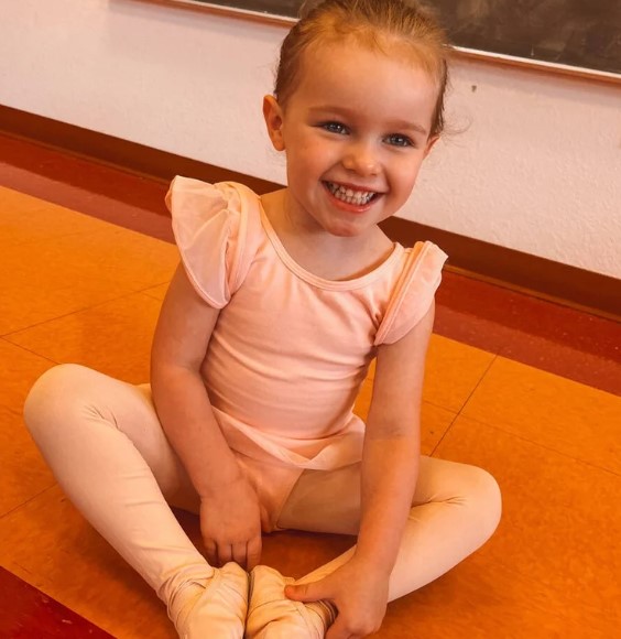 Little girl in a peach leotard and tights sits on the studio floor with her legs crossed